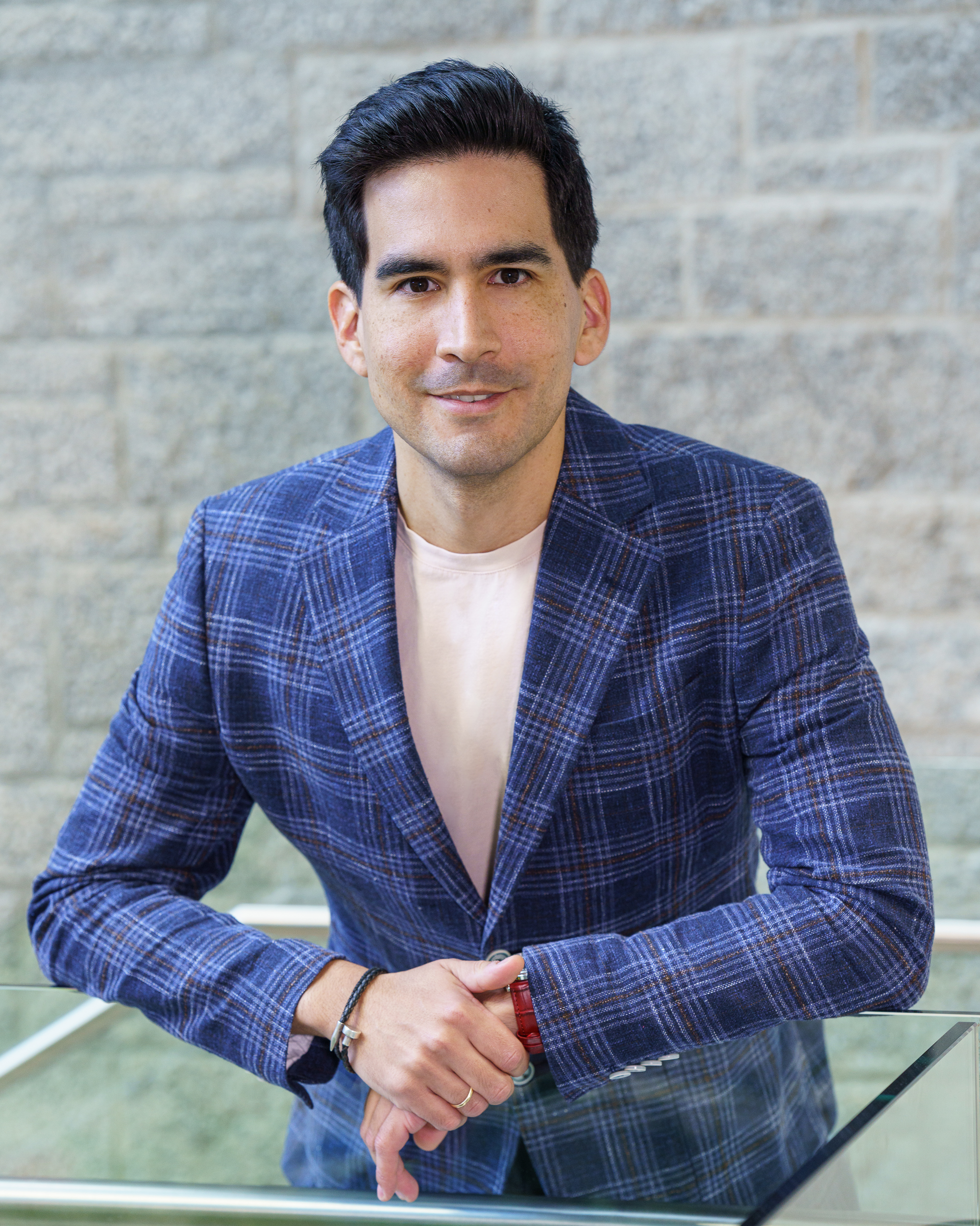 A man with short dark hair wearing a blue plaid blazer over a light shirt leans forward, resting his hands on a glass surface, with a stone wall in the background.