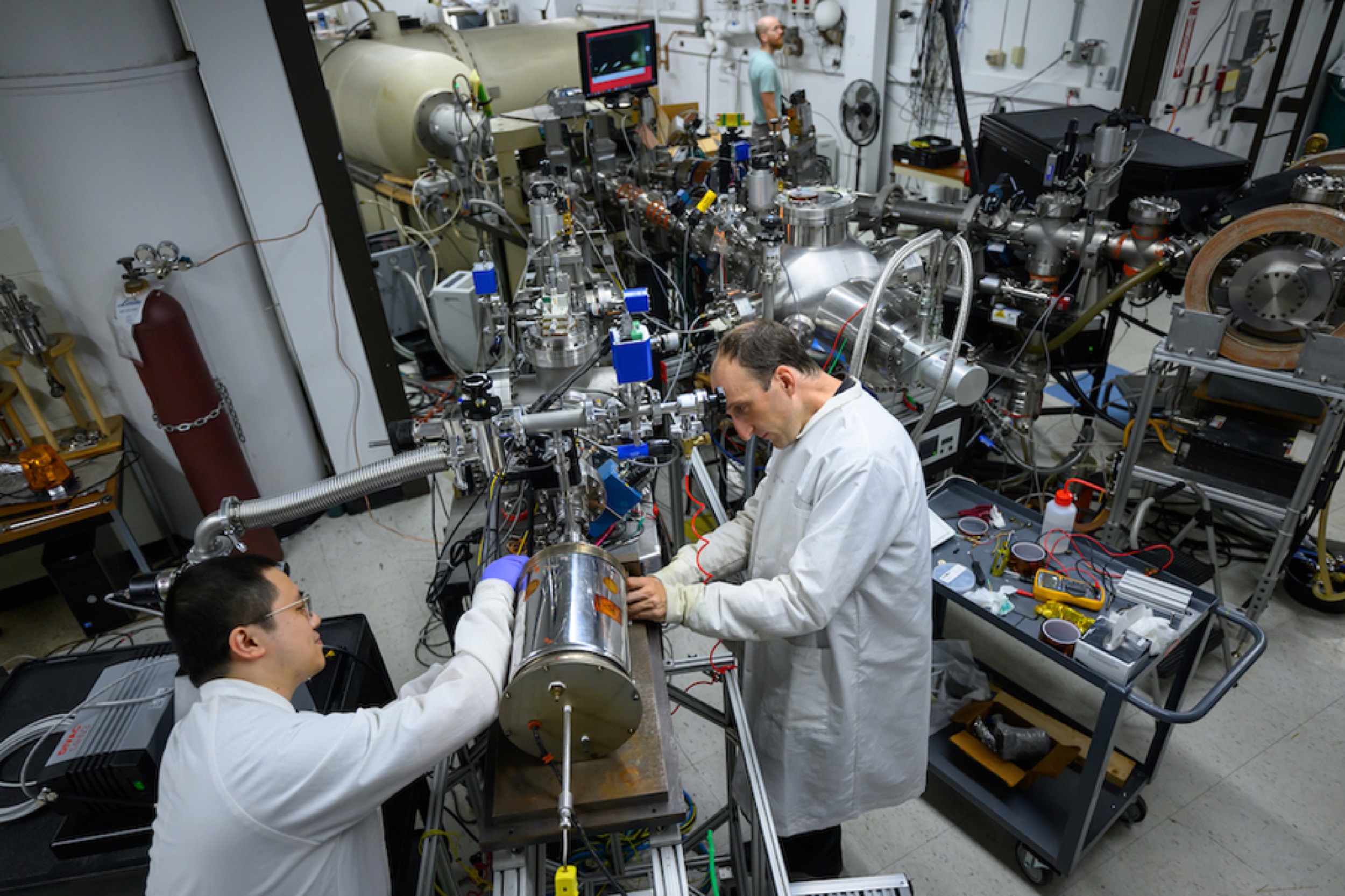 Two scientists in lab coats work with complex instruments and metal equipment in a busy laboratory. The room contains various machines, cables, and scientific devices, with another person seen in the background.