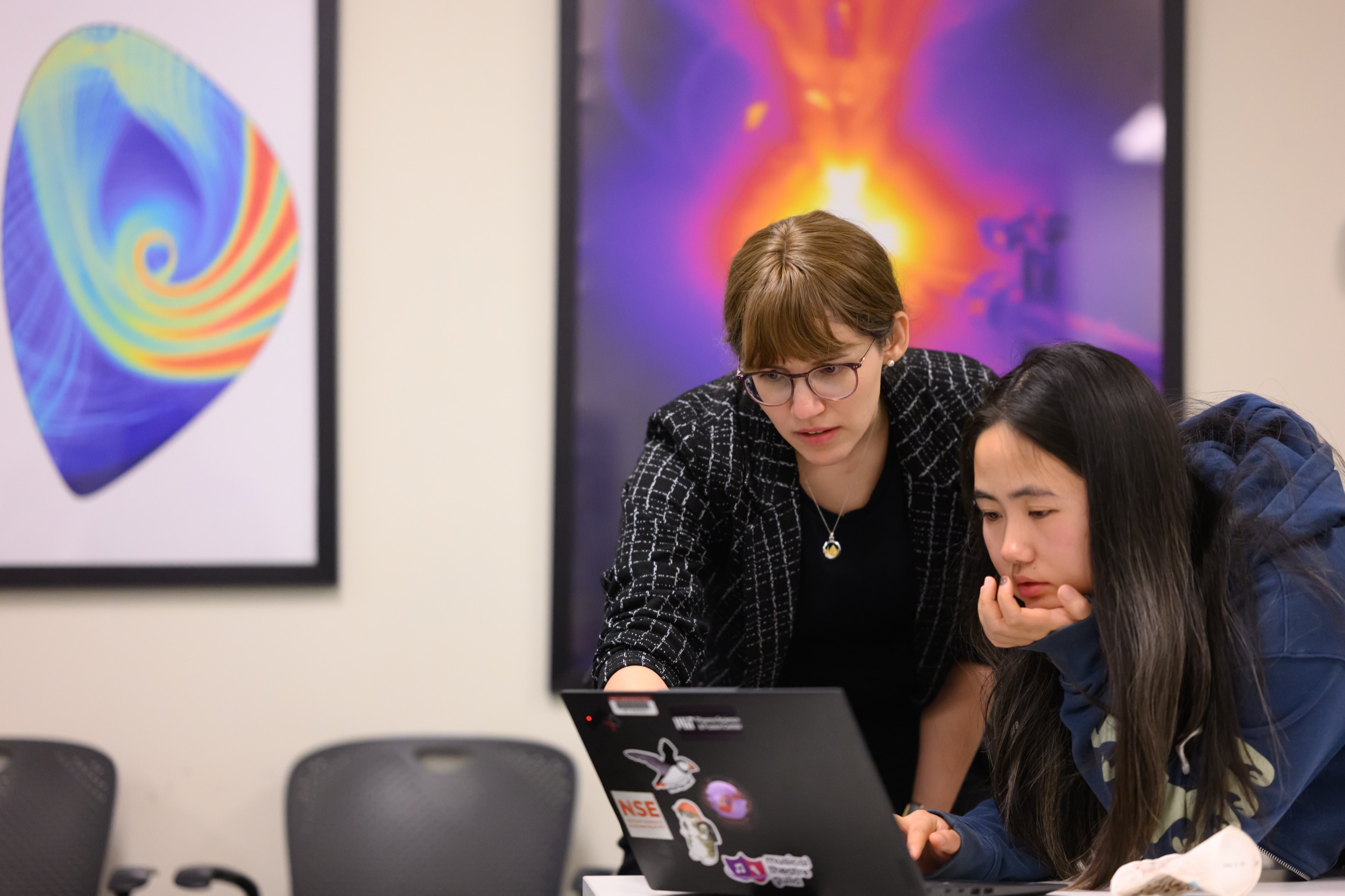 Two women look intently at a laptop screen, one standing and pointing while the other sits, resting her chin on her hand. Colorful abstract art is visible on the wall behind them.