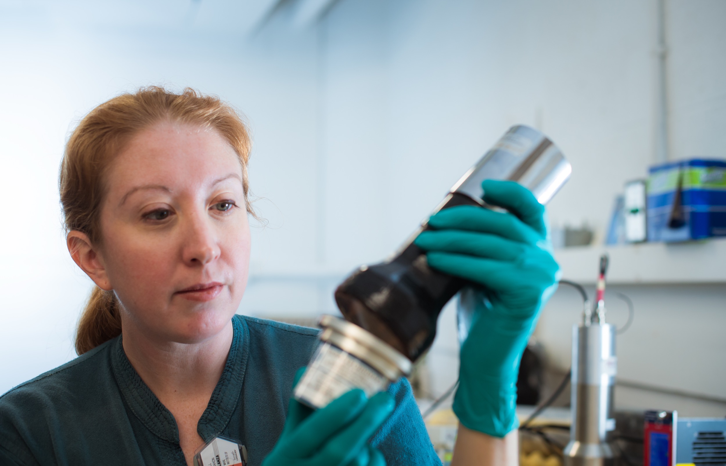A woman wearing teal gloves examines a laboratory container with a focused expression in a bright lab setting. Shelves and scientific equipment are visible in the background.