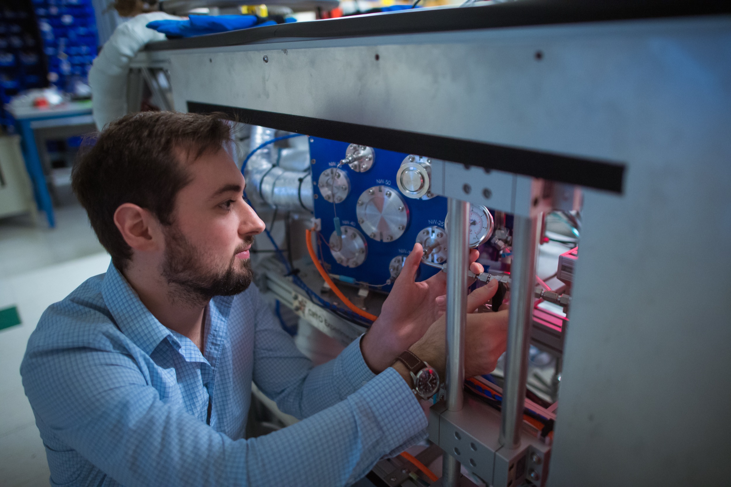 A man in a light blue shirt works on a complex machine with wires and metal components in a laboratory setting, adjusting parts with his hands.