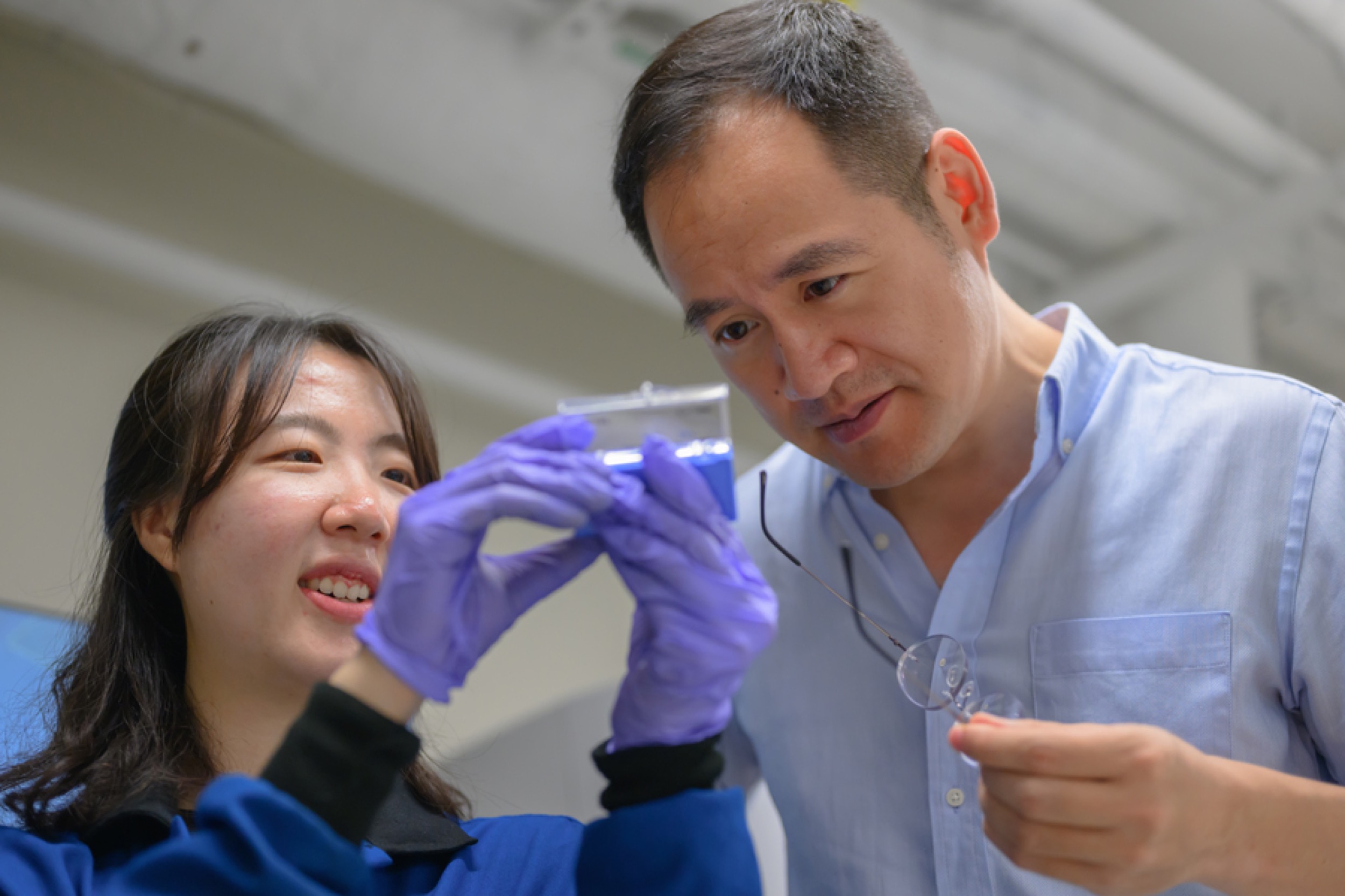 A woman wearing purple gloves shows a transparent container to a man in a light blue shirt, who holds his glasses and looks at the object closely. They appear to be in a lab setting.