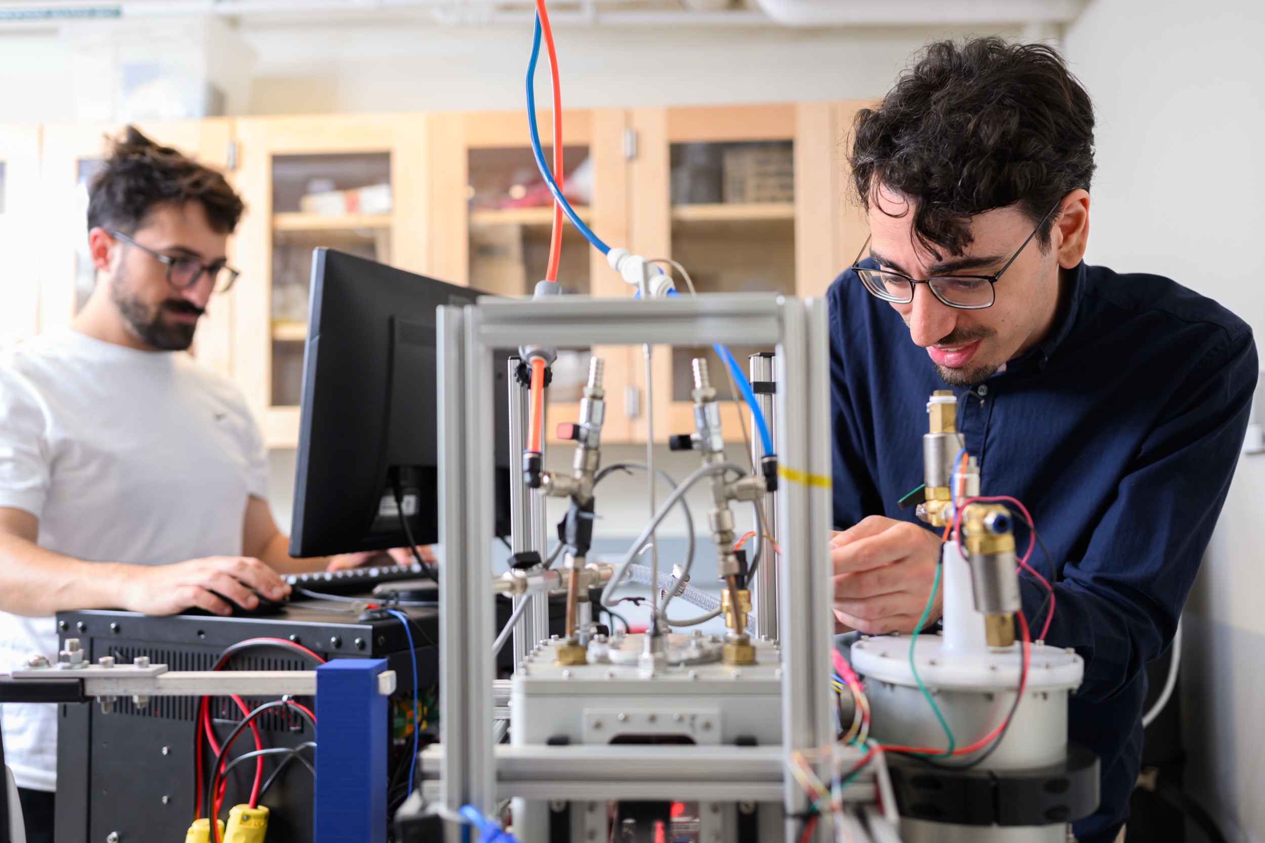Two men in a laboratory work with electronic equipment and wires; one is adjusting a device, while the other uses a computer. The workspace has shelves and various technical components visible.