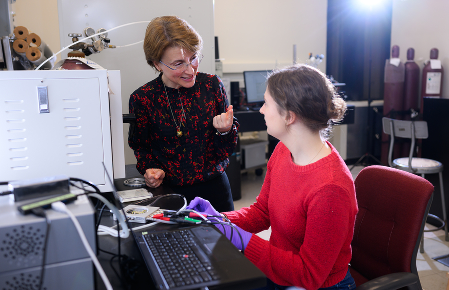 A woman in a black floral blouse speaks to a young woman in a red sweater and purple gloves, who is seated at a computer workstation in a laboratory filled with scientific equipment.