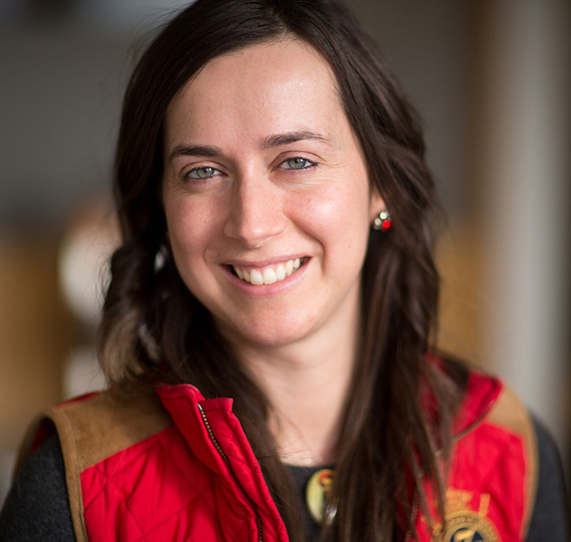 A woman with long brown hair smiles at the camera. She is wearing a red vest over a dark top, and small red earrings. The background is softly blurred.