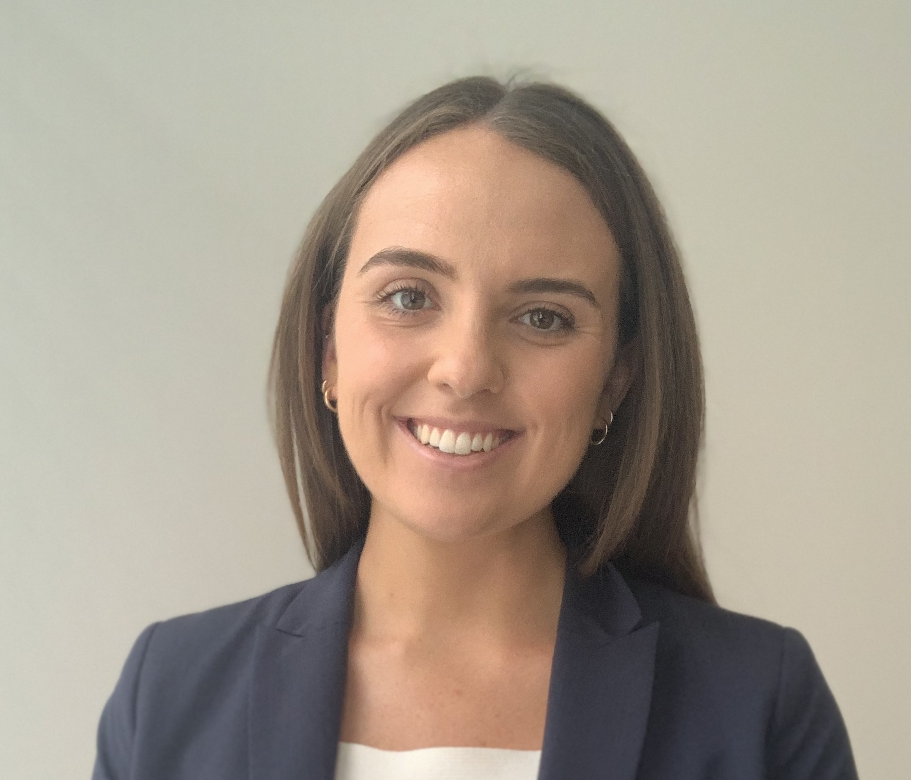 A woman with straight brown hair, wearing a navy blazer and white top, smiles at the camera against a plain light background.