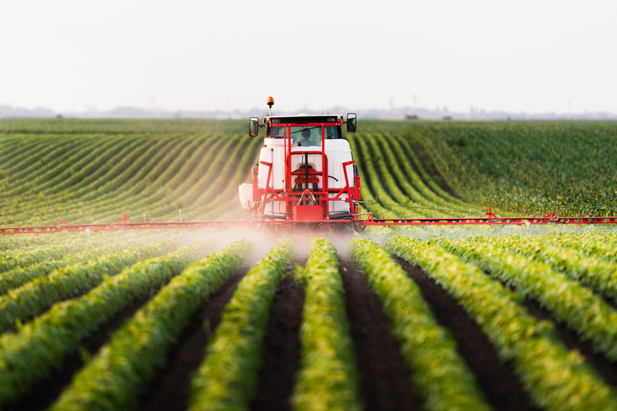 A red agricultural machine sprays crops in a green, neatly lined field, likely applying fertilizer or pesticides under a cloudy sky.