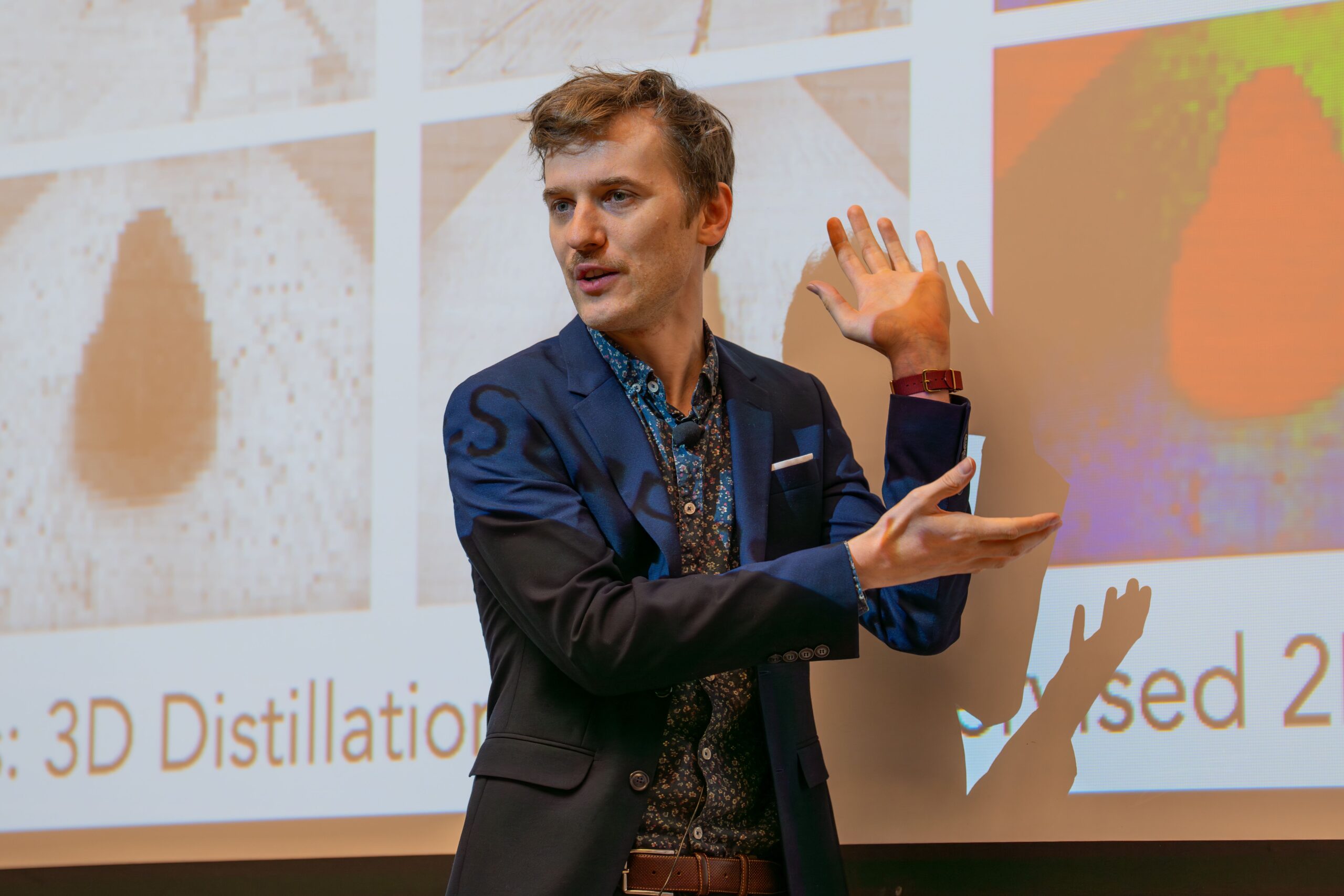 A man in a navy suit gestures with both hands while presenting in front of a screen displaying scientific images and partial text.