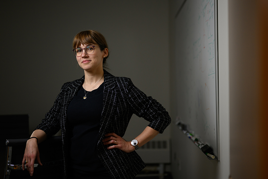 A woman wearing glasses and a dark outfit stands confidently with one hand on her hip in a dimly lit office next to a whiteboard with faint writing.