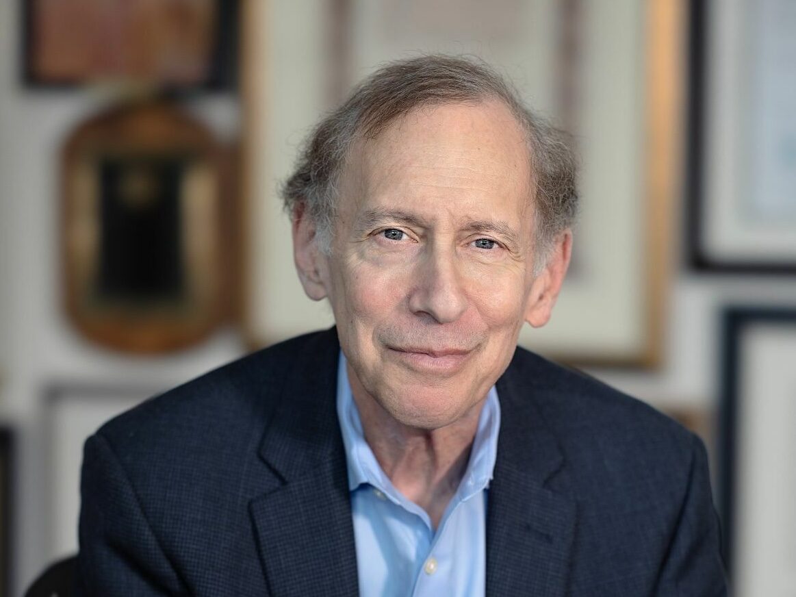 A middle-aged man with thinning hair, wearing a blue dress shirt and dark blazer, sits and smiles softly. The background features blurred framed certificates and awards.