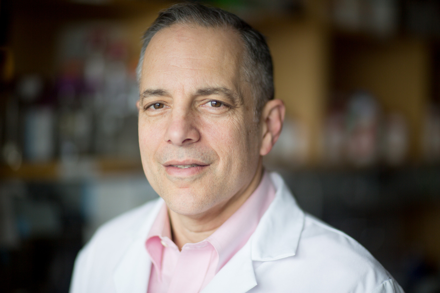 A middle-aged man with short gray hair wearing a white lab coat and a light pink shirt stands in a blurred laboratory setting, looking at the camera with a neutral expression.