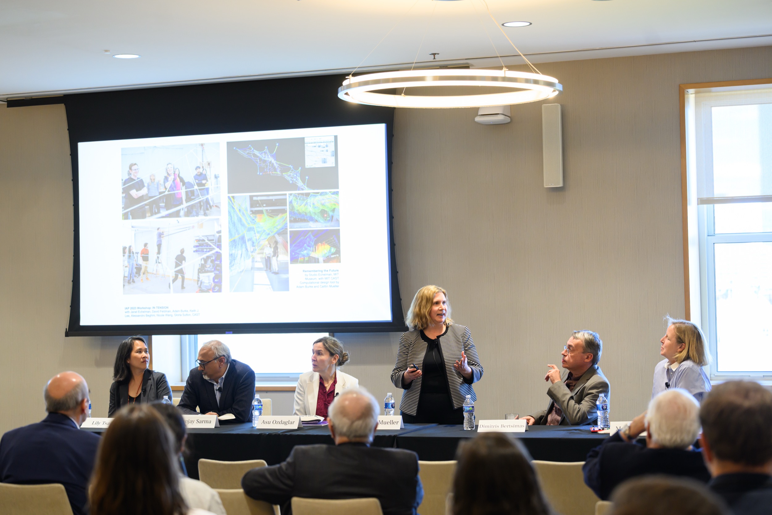 A woman stands and speaks at a panel discussion with six seated panelists in front of an audience. A large screen behind them displays scientific images and diagrams.