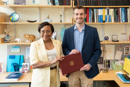 Two people stand in an office, smiling at the camera. The woman on the left is wearing a light-colored suit, and the man on the right, wearing a blue blazer, holds a red certificate folder. Books and photos fill the shelves behind them.