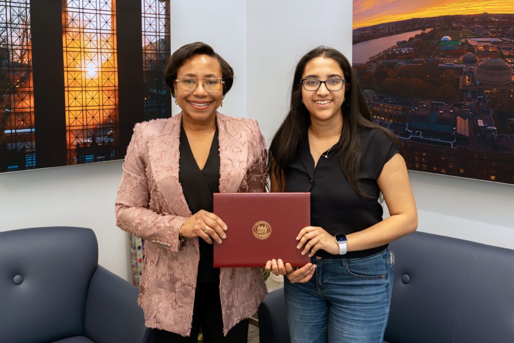 Two women stand indoors, smiling at the camera. One woman, wearing a light pink blazer, and the other, in a black shirt and jeans, hold a maroon certificate folder together. Framed photos of cityscapes hang behind them.