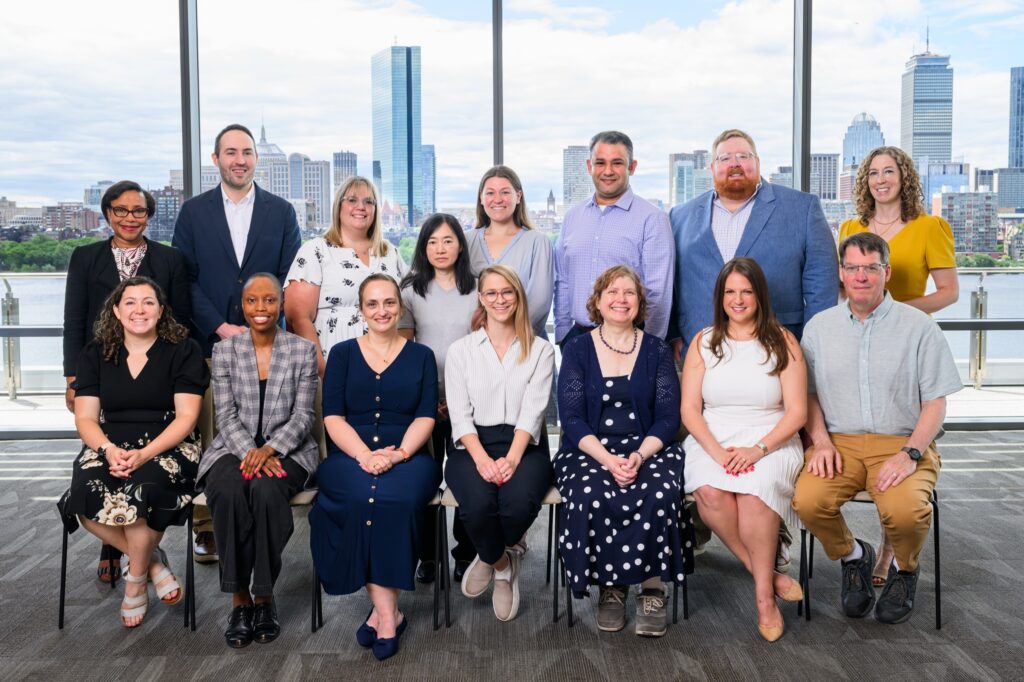 A group of fifteen adults pose indoors in front of large windows with a city skyline and river in the background. They are dressed in business casual attire, and all are smiling at the camera.