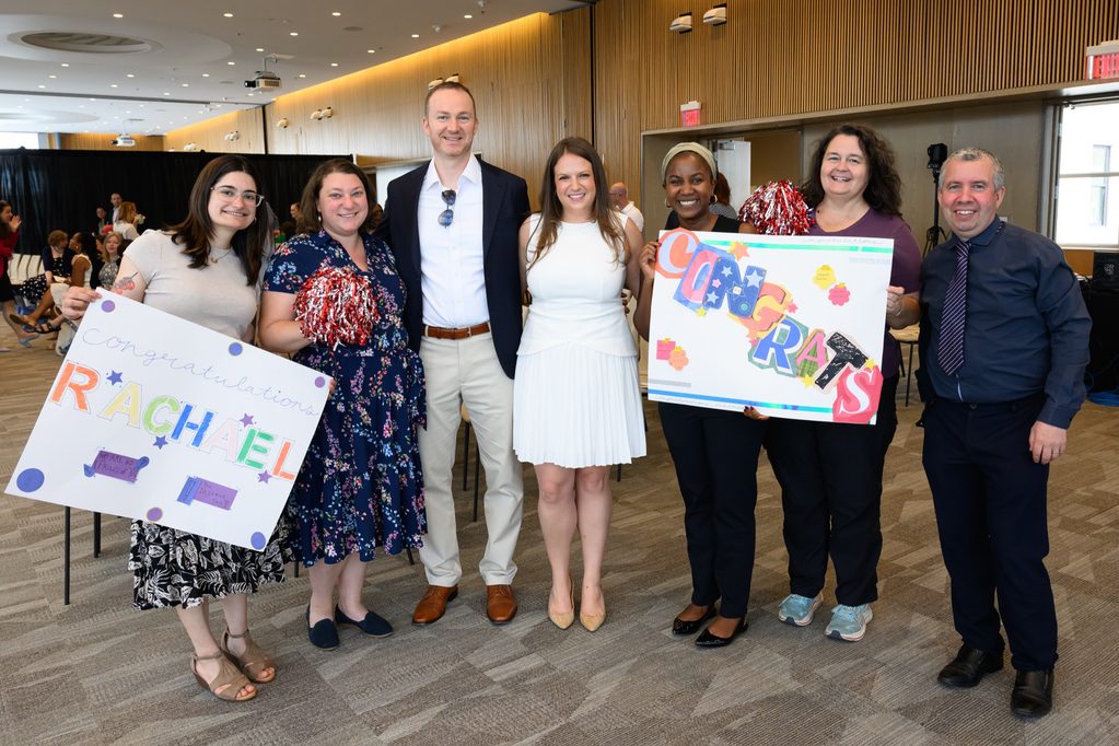 Seven people smile at an indoor event. Two hold handmade signs: one says Congratulations Rachel, and another says Congrats. The group stands together, posing happily for the photo.