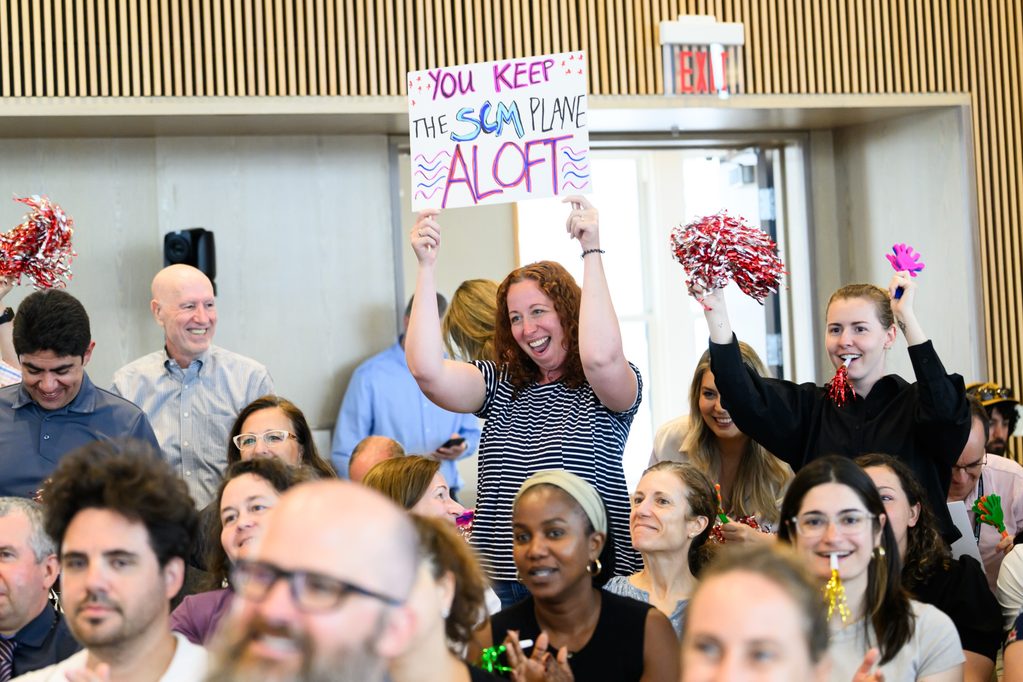 A smiling woman stands among a seated crowd, holding a colorful sign that reads You keep the SCM plane aloft, while others around her cheer and clap enthusiastically.