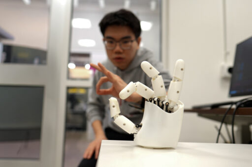 A person makes an OK hand gesture toward a white robotic hand, which mimics the same gesture, in a modern indoor setting.