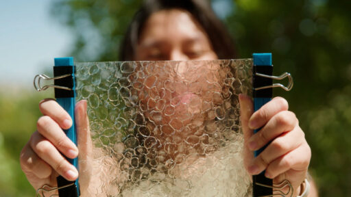 A person holds a rectangular, transparent sheet with a bubbly texture, secured by binder clips on each side. Green trees and sky are blurred in the background.