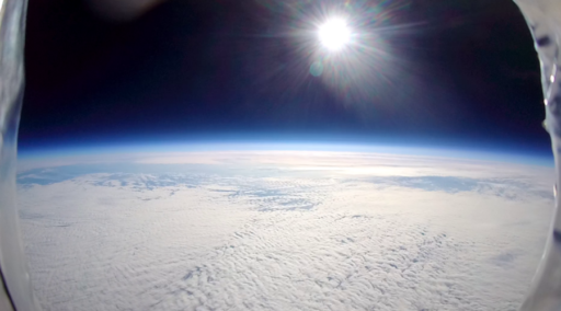The sun shines brightly over Earths curved horizon, with a blanket of clouds below and the dark edge of space above.