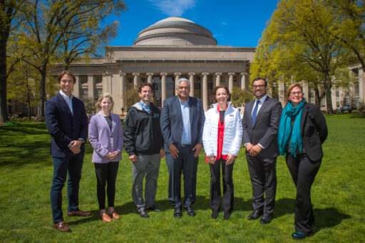 Seven people stand in a row on a grassy lawn in front of a large domed building with columns, likely on a university campus, with bright sunlight and trees in the background.
