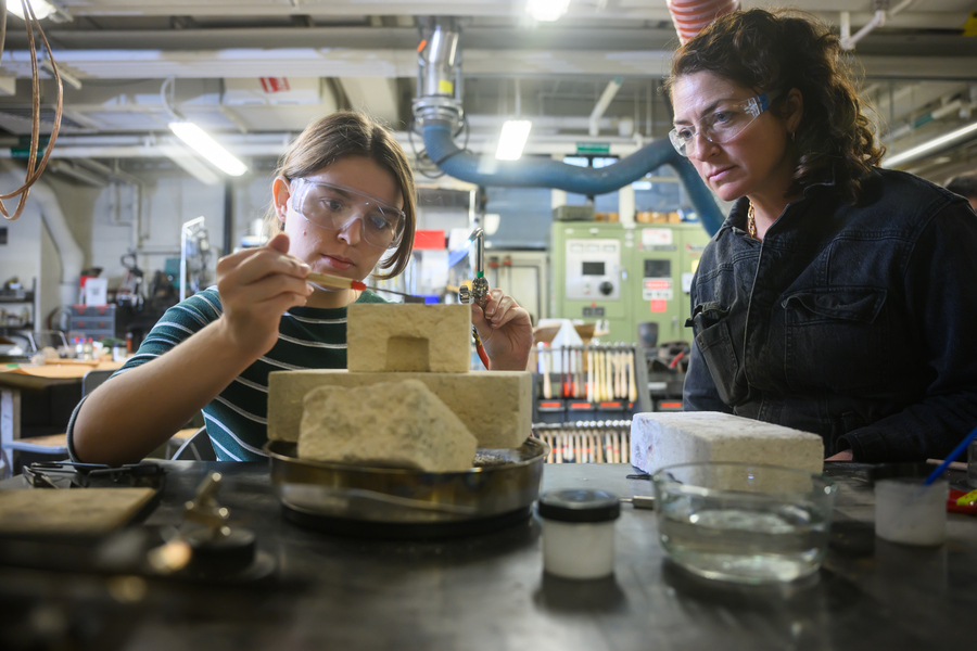 Two women wearing safety glasses work on a project in a workshop, using tools and stone blocks on a workbench, surrounded by industrial equipment and supplies.