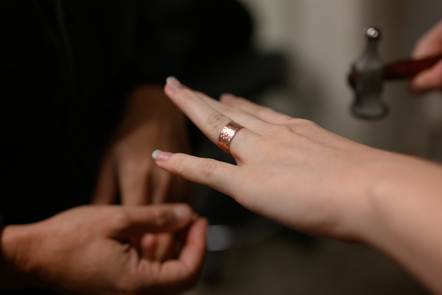 A person wears a textured copper ring on their finger while another hand holding a small hammer approaches, possibly to adjust the ring. Another person’s hands are nearby, slightly out of focus.